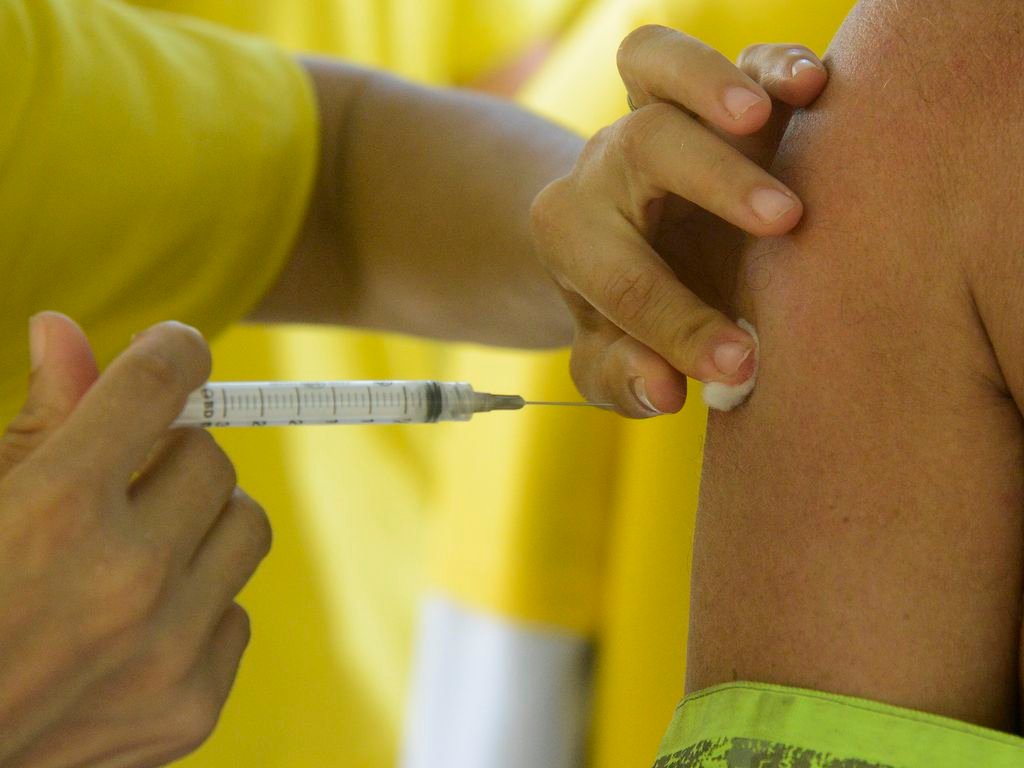 Gripe #PraCegoVer Na foto, uma pessoa com camisa amarela aplica uma injeção com vacina contra a gripe em outra pessoa