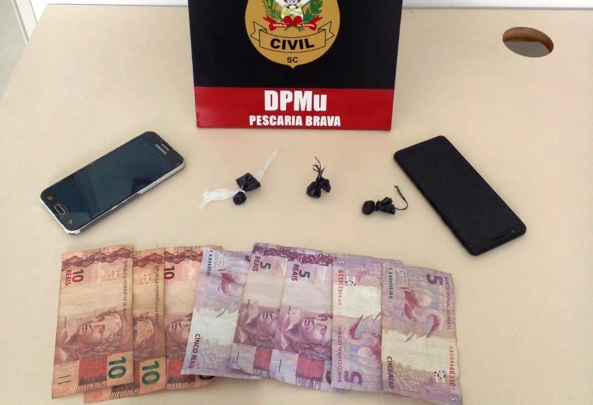 operacao-pc-pescaria-brava #ParaTodosVerem Na foto, uma placa com o símbolo da Polícia Civil, dois aparelhos de telefone celular, algumas cédulas de dinheiro e três porções de drogas