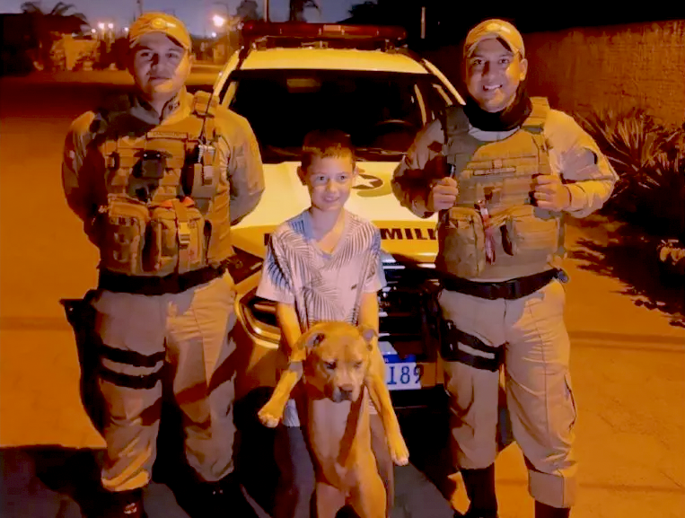 PM Navegantes #ParaTodosVerem Na foto, há dois policiais militares, um criança e um cachorro que foi furtado e encontrado. No fundo, há a viatura da PM