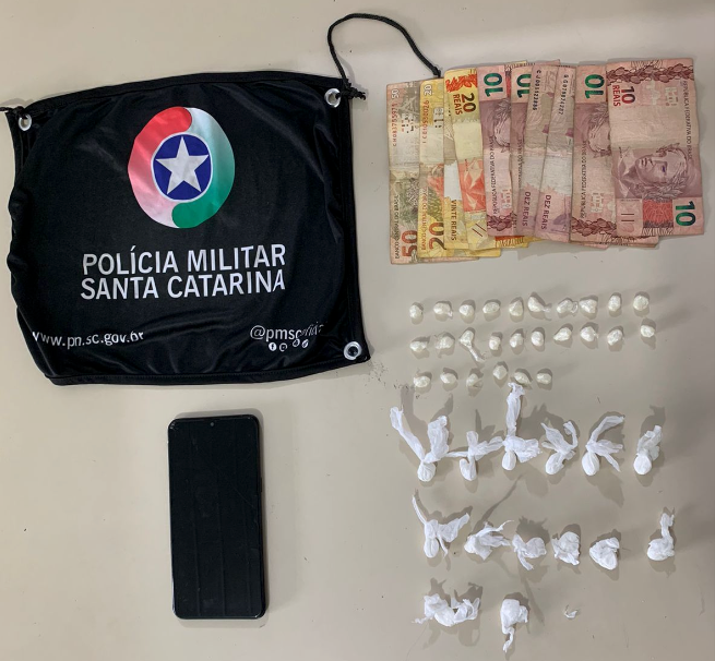 Tráfico TB #ParaTodosVerem Na foto, sobre uma mesa, há uma bandeirola com o símbolo da Polícia Militar de Santa Catarina, cédulas de dinheiro, porções de drogas e um aparelho de telefone celular preto