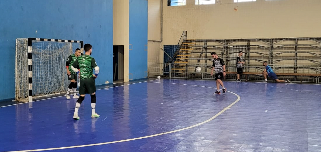 Tubarão Futsal 1 #ParaTodosVerem Na foto, três atletas do Tubarão Futsal treinam chutes. No fundo, outros dois atletas do time fazem alongamentos