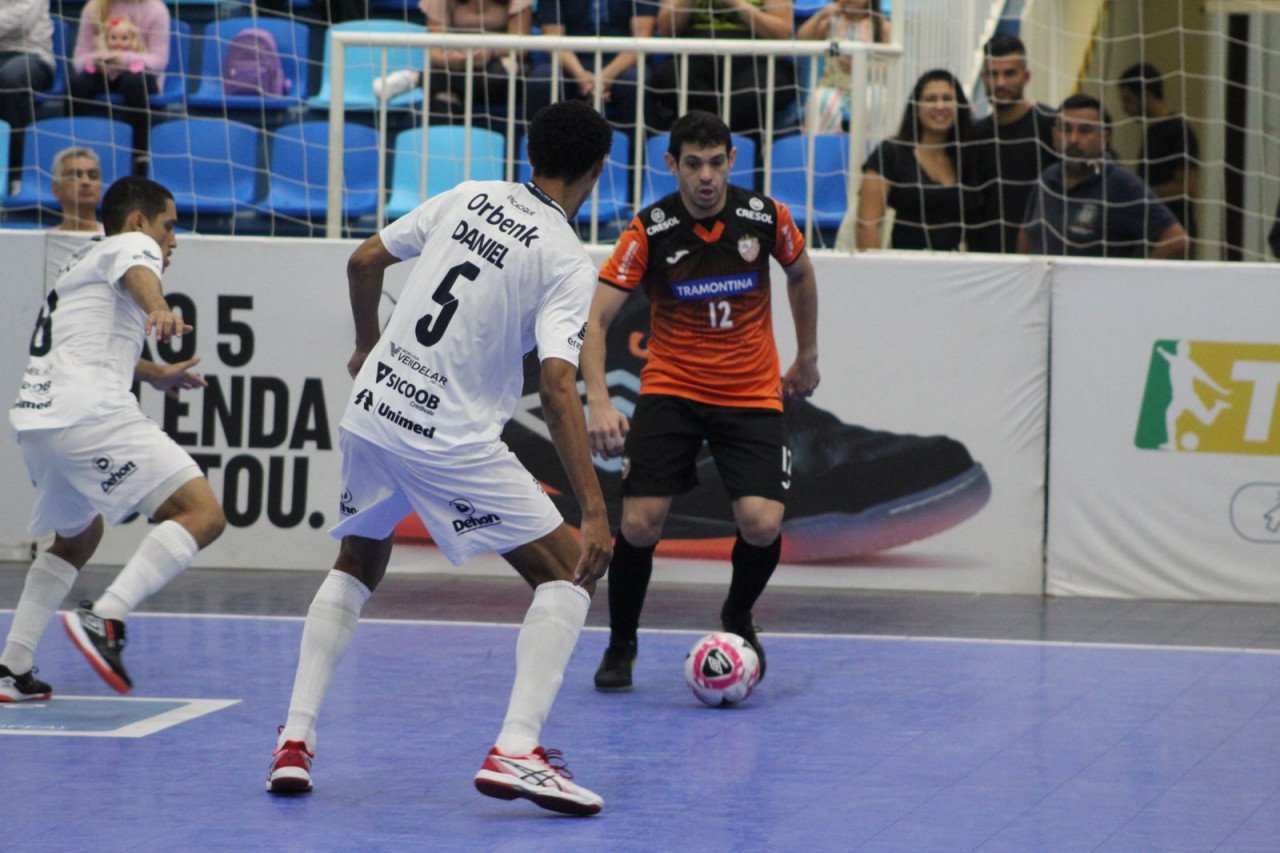 Tubarão Futsal #PraCegoVer Na foto, dois jogadores de futsal com uniforme branco e um atleta com traje laranja e preto disputam a posse de bola
