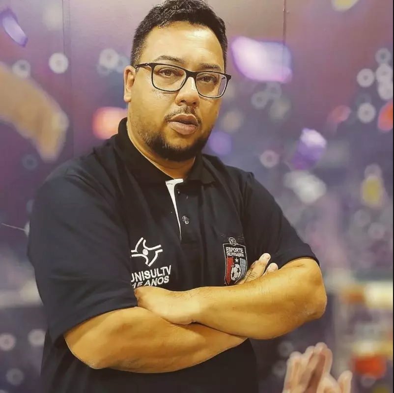 Ventura #ParaTodosVerem Na foto, o jornalista Eduardo Ventura, um homem moreno e que uso óculos. Ele aparece em um fundo desfocado e está de camiseta preta e braços cruzados