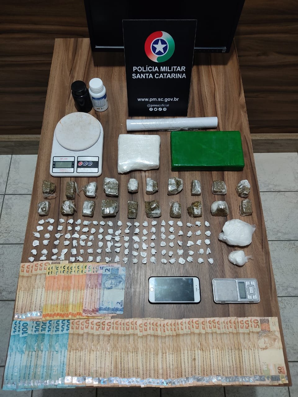 Droga Imbituba #ParaTodosVerem Na foto, em cima de uma mesa há diversas notas de dinheiro, porções de cocaína e maconha, balanças de precisão, um aparelho de telefone celular e filme plástico utilizado para porcionamento das drogas