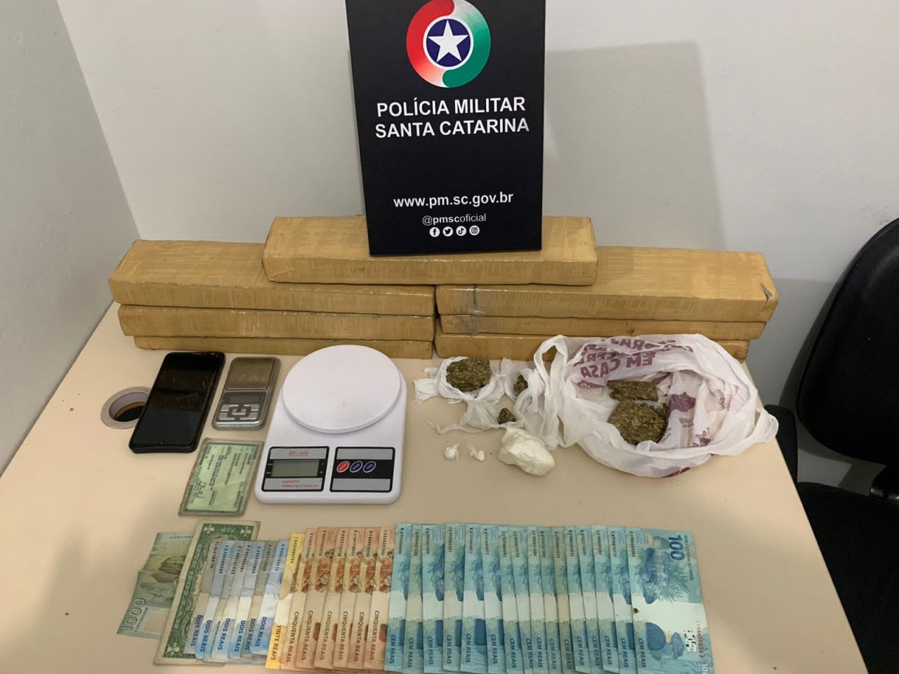 Droga #ParaTodosVerem Na foto, em cima de uma mesa há uma placa com o símbolo da Polícia Militar em cima de tabletes grandes de maconha, porções menores de cocaína e maconha, cédulas de dinheiro brasileiro e estrangeiro, duas balanças de precisão, um aparelho de telefone celular preto