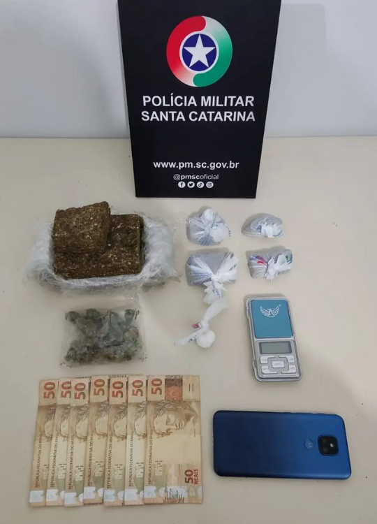 Drogas Imbituba #ParaTodosVerem Na foto, em cima de uma mesa há uma placa com o símbolo da Polícia Militar de Santa Catarina, porções de maconha e cocaína, uma balança de precisão, dinheiro e um aparelho de telefone celular azul