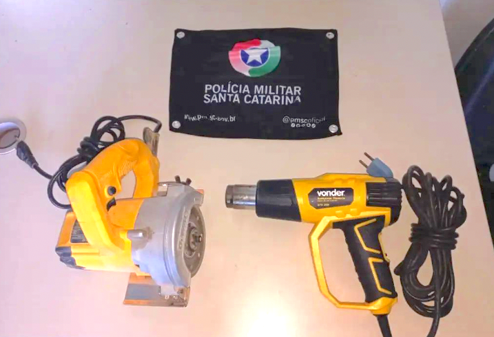 Maquinas furtadas imbituba #ParaTodosVerem Na foto, em cima de uma mesa há uma bandeira com o símbolo da Polícia Militar, uma maquina e um soprador térmico