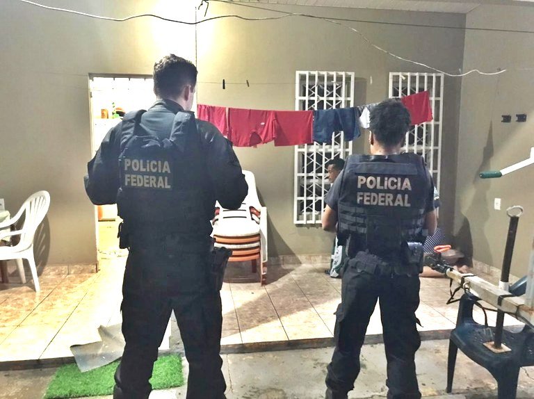 Operação ODisseu #ParaTodosVerem Na foto, dois policiais federais estão de costas em frente de uma casa, em Roraima, onde cumprem um mandado de busca e apreensão