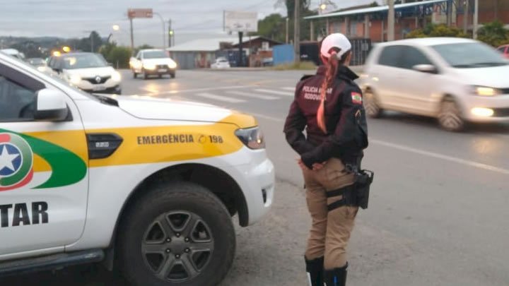 PMRv - cópia #ParaTodosVerem Na foto, uma policial militar rodoviária está ao lado de uma viatura, às margens de uma estrada, fiscalizando o trânsito