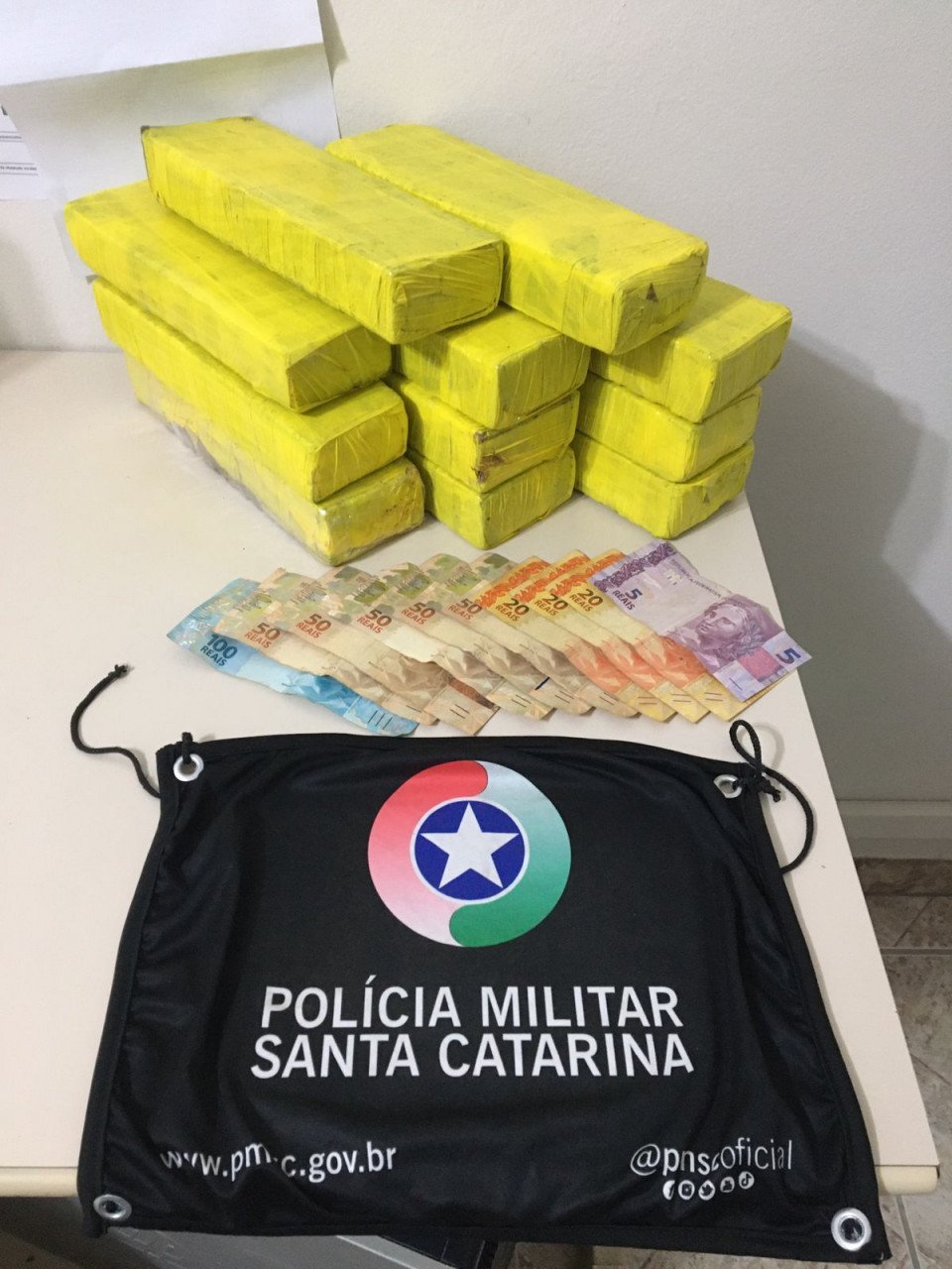 Maconha Tubarão #ParaTodosVerem Na foto, em cima de uma mesa há 11 tabletes amarelos de maconha, cédulas de dinheiro e uma bandeira com o símbolo da Polícia Militar