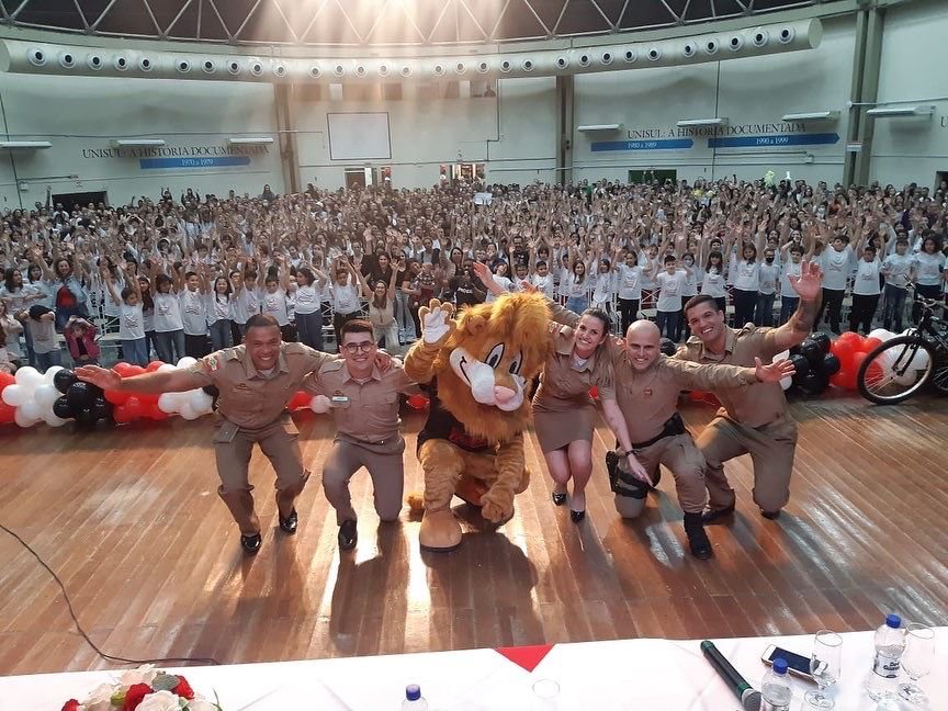 Proerd #ParaTodosVerem Na foto, em cima do palco, estão os policiais militares responsáveis por desenvolver o Proerd em Tubarão e o leão Daren, o mascote da ação. No fundo, os 865 alunos diplomados nesta semana junto com seus pais e professores