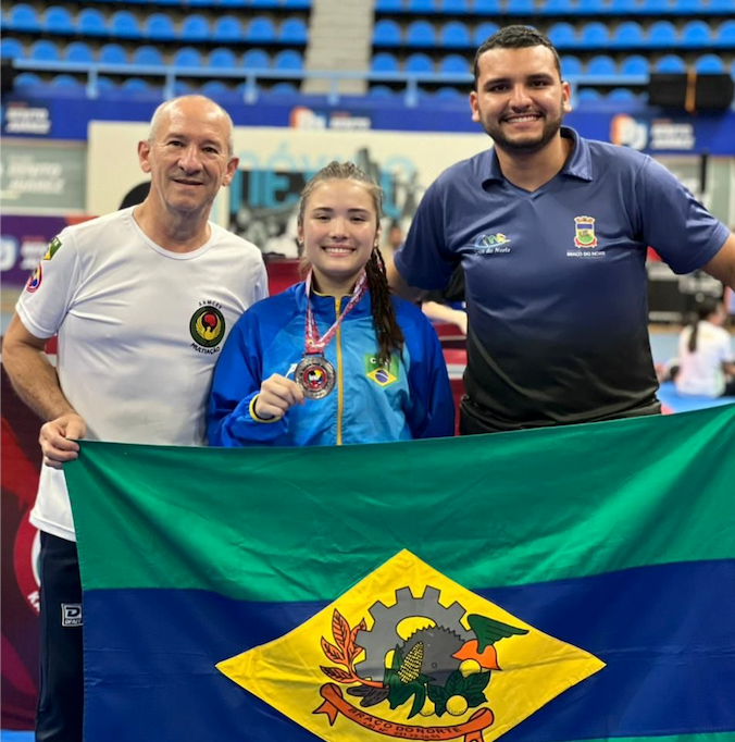 Captura de Tela 2022-08-26 às 17.40.19 #ParaTodosVerem Na foto, dois homens e uma garota seguram a bandeira do município de Braço do Norte