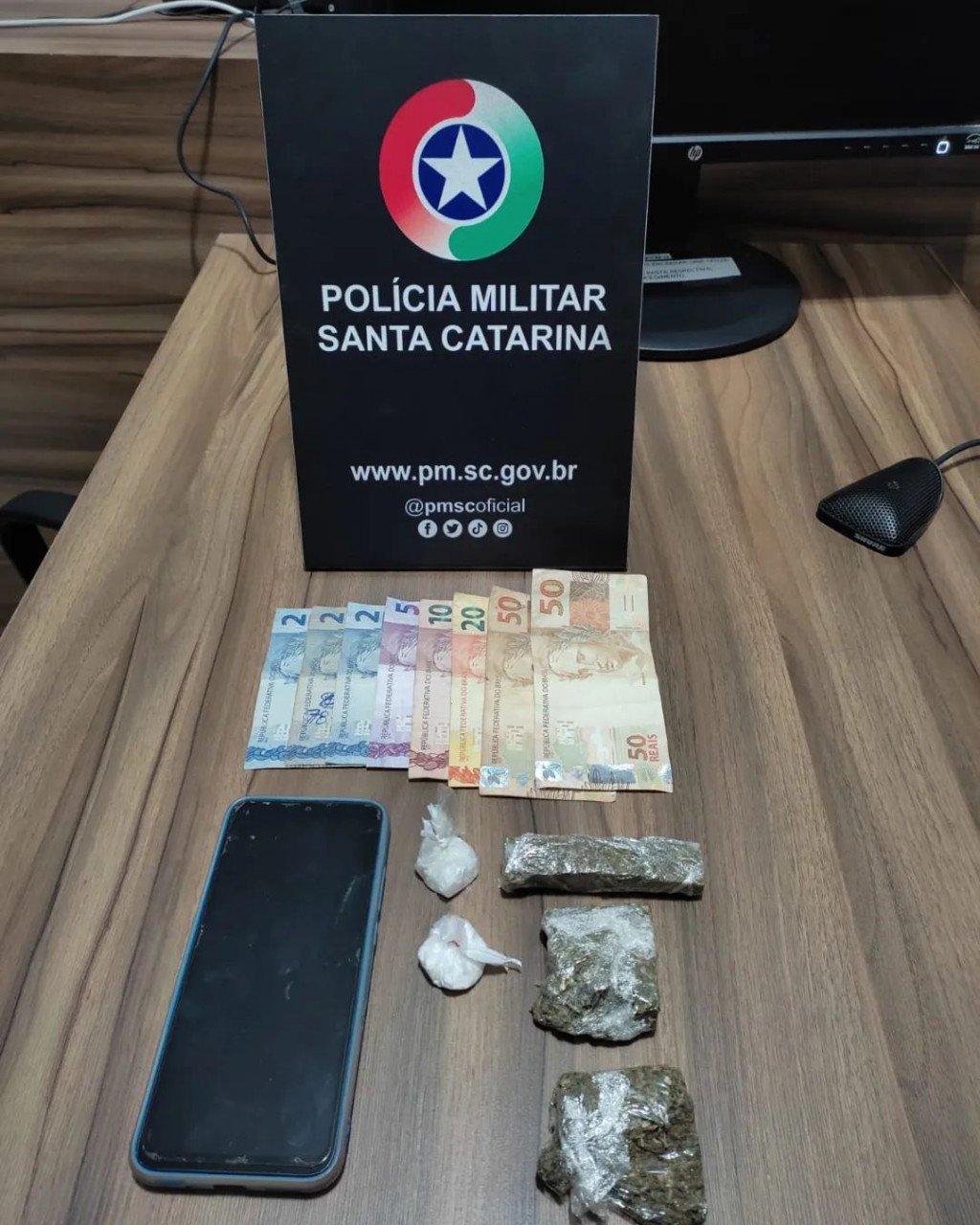 pmscimbituba34bpm_299850285_113616828123229_8276789480722046027_n #ParaTodosVerem Na foto, em cima de uma mesa há uma placa com o símbolo da Polícia Militar, porções de drogas, um telefone celular preto e algumas cédulas de dinheiro