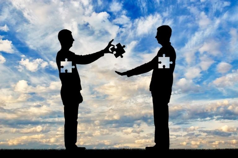 #ParaTodosVerem Na foto, um céu azul com nuvens brancas. Na frente, duas silhuetas masculinas. Uma entrega uma peça de quebra-cabeça para a outra