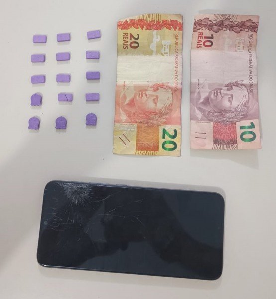 tubarao-adolescente-e-detido-com-15-comprimidos-de-ecstasy #ParaTodosVerem Na foto, comprimidos de ecstasy, um aparelho de telefone celular preto com a tela danificada e alguns cédulas de dinheiro